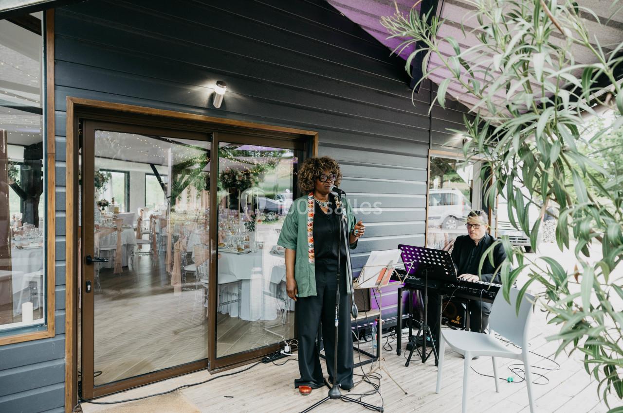 Une chanteuse se produit sur une terrasse en bois, accompagnée d'un pianiste, devant une salle vitrée décorée.