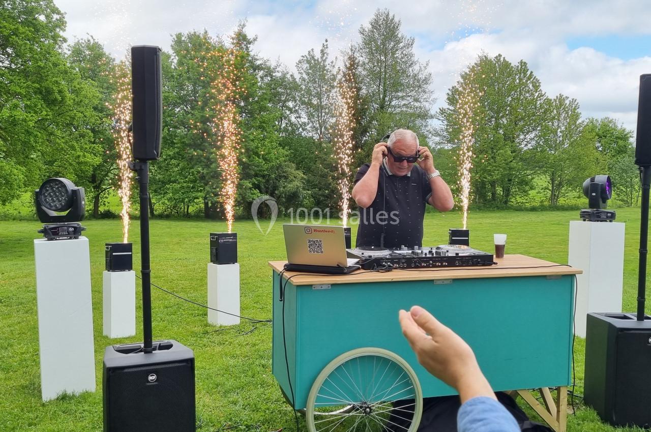 Un DJ mixe en plein air sur une table mobile, entouré d'effets pyrotechniques et de haut-parleurs dans un parc verdoyant.