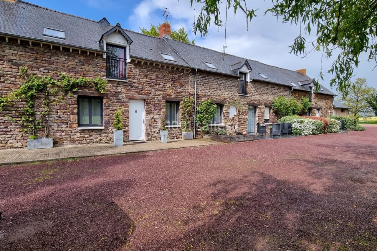 Façade d'une maison en pierre avec toit en ardoise, entourée de végétation et donnant sur une cour en gravier rouge.
