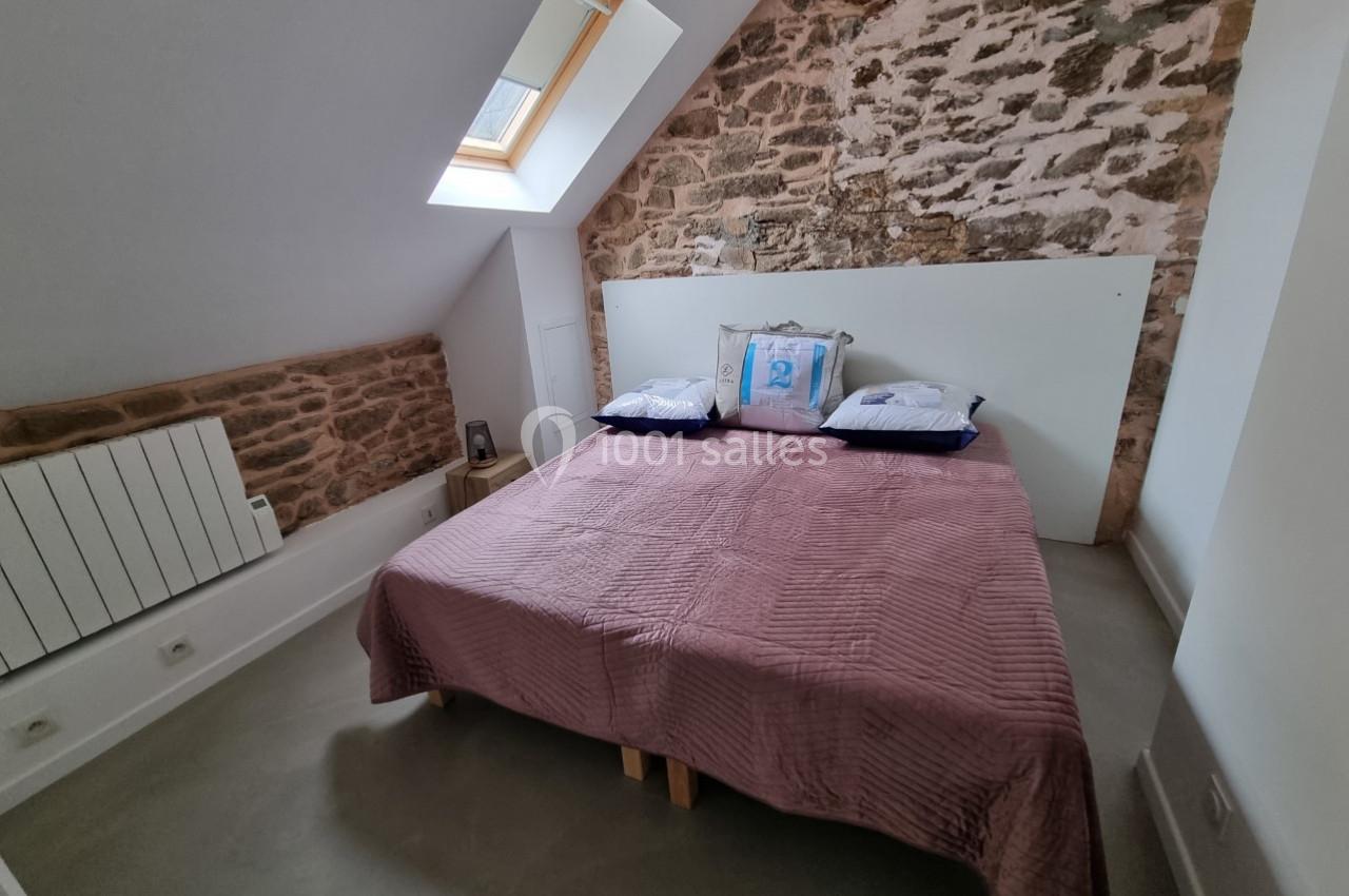 Chambre mansardée avec un lit double, mur en pierres apparentes, fenêtre de toit et sol en béton lisse.