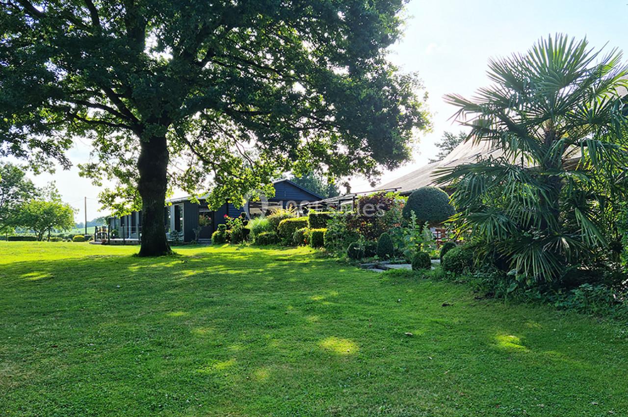 Jardin verdoyant avec pelouse, grands arbres et arbustes, bordant des bâtiments en arrière-plan sous un ciel clair.