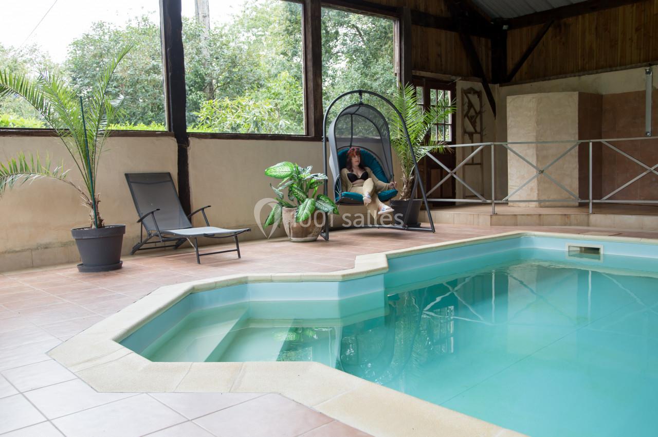 Piscine intérieure avec carrelage beige, transat, plantes en pot et une personne assise dans une chaise suspendue.
