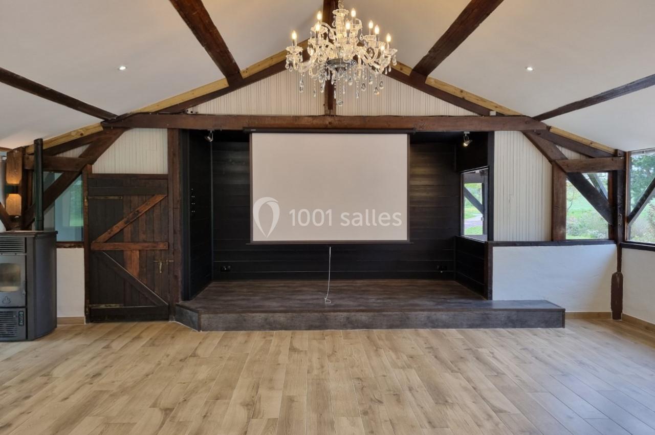 Salle avec parquet, scène en bois sombre, écran blanc, poutres apparentes et lustre central.
