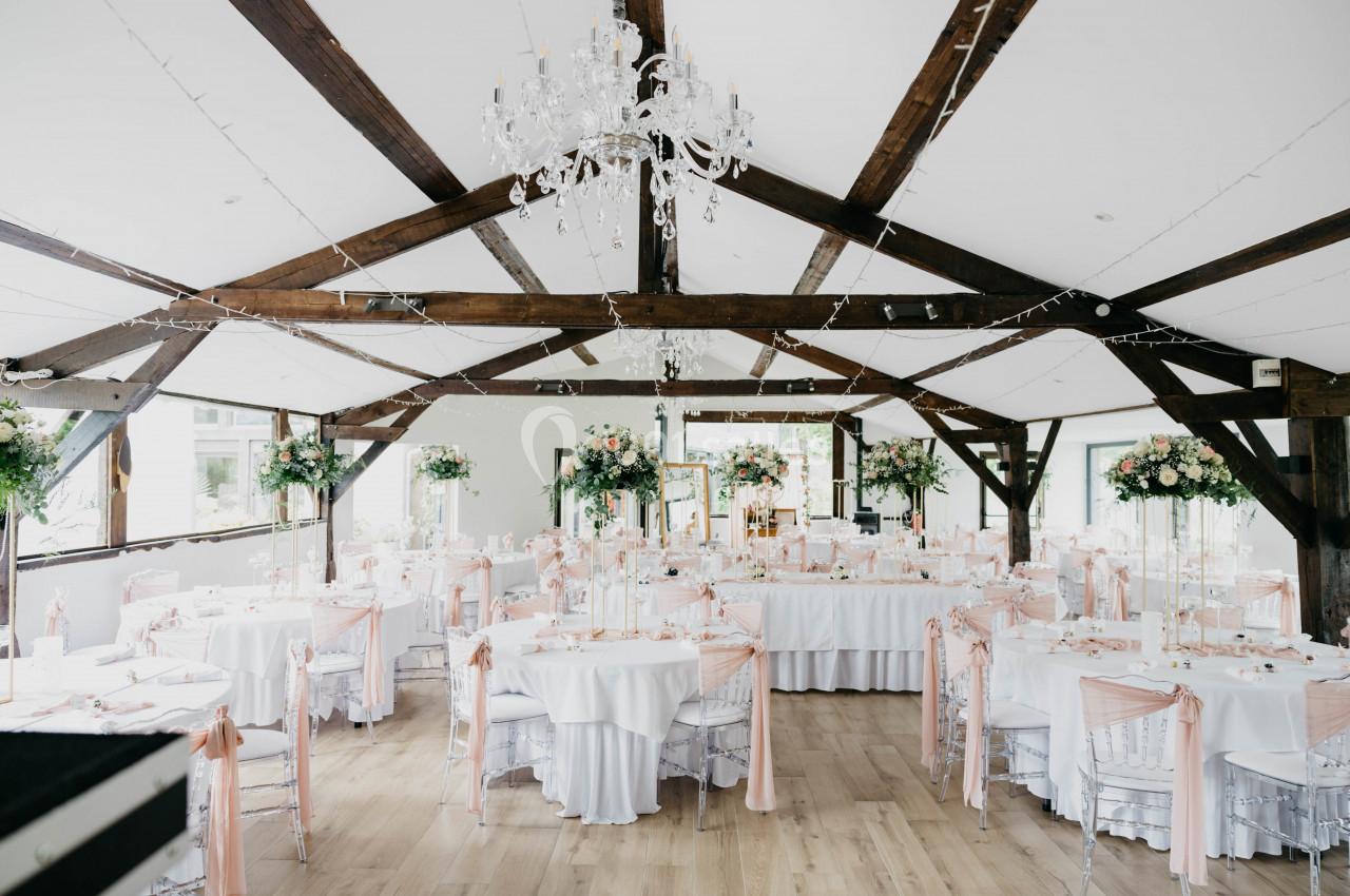 Salle de réception lumineuse avec tables rondes décorées de nappes blanches, rubans roses et centres de table fleuris.