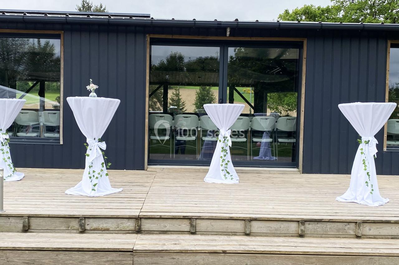 Tables hautes décorées de nappes blanches et de feuillages, disposées sur une terrasse en bois devant un bâtiment vitré.