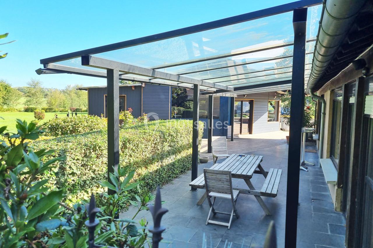 Terrasse extérieure avec pergola en verre, mobilier en bois et vue sur un jardin verdoyant par temps ensoleillé.