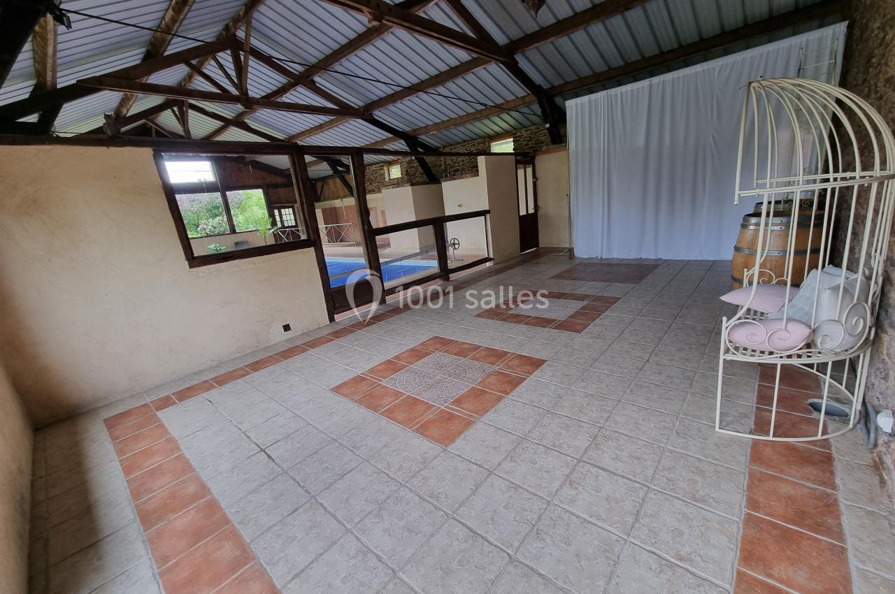 Salle carrelée avec toit en tôle, grandes ouvertures vitrées donnant sur une piscine, rideau blanc et étagère métallique.