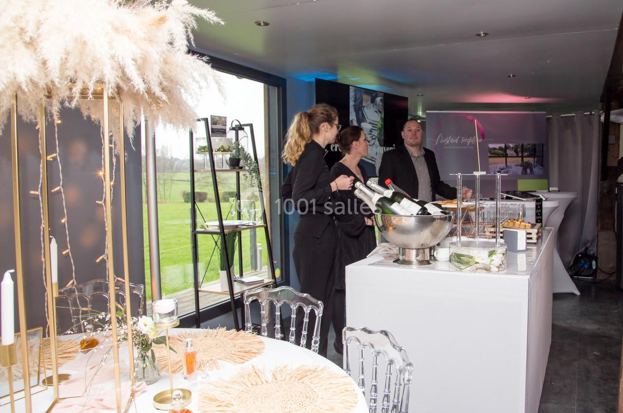 Trois personnes discutent près d'un bar avec des bouteilles de champagne, dans une salle décorée pour un événement.