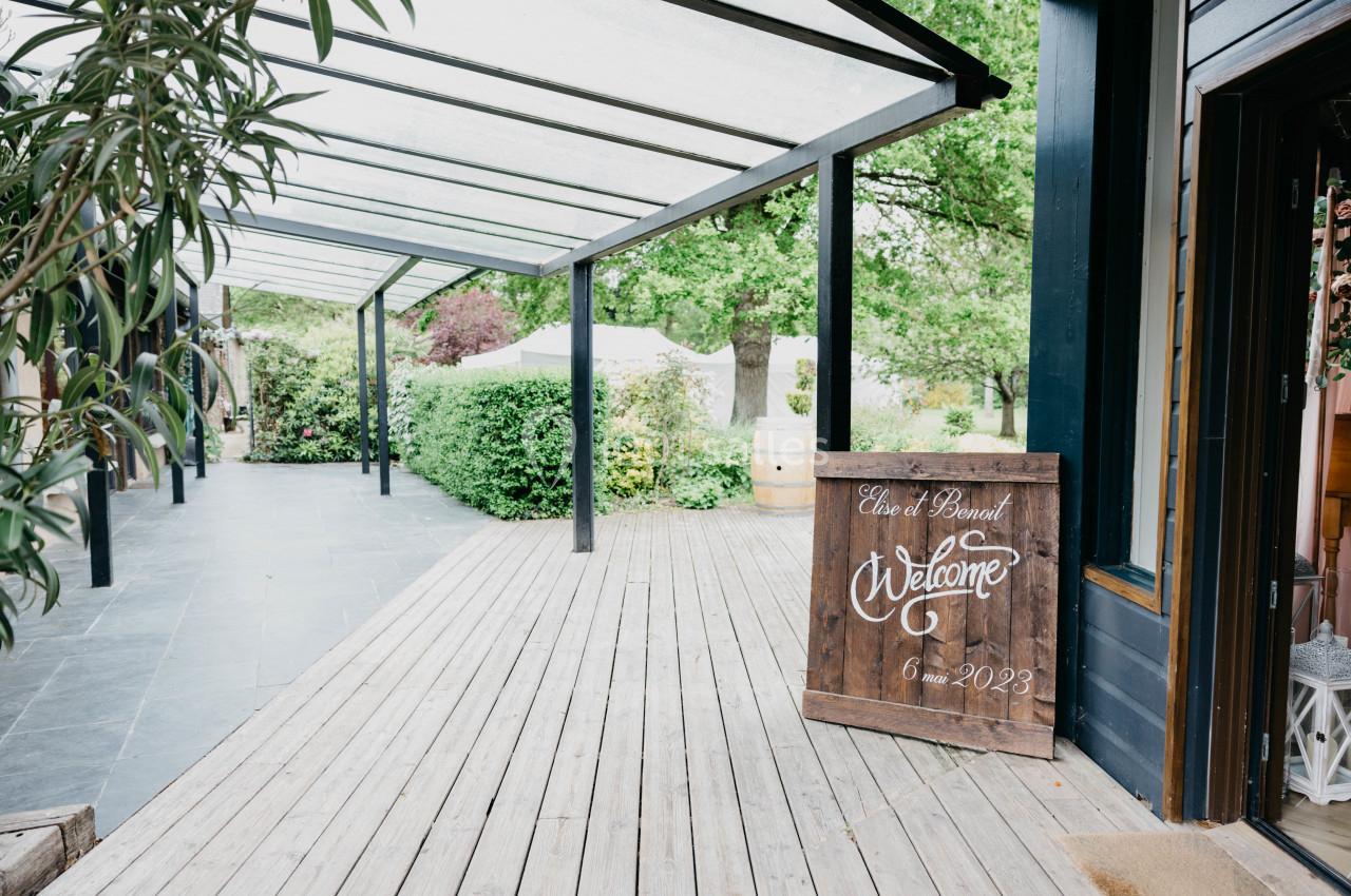 Terrasse en bois couverte, entourée de verdure, avec un panneau de bienvenue indiquant une date.