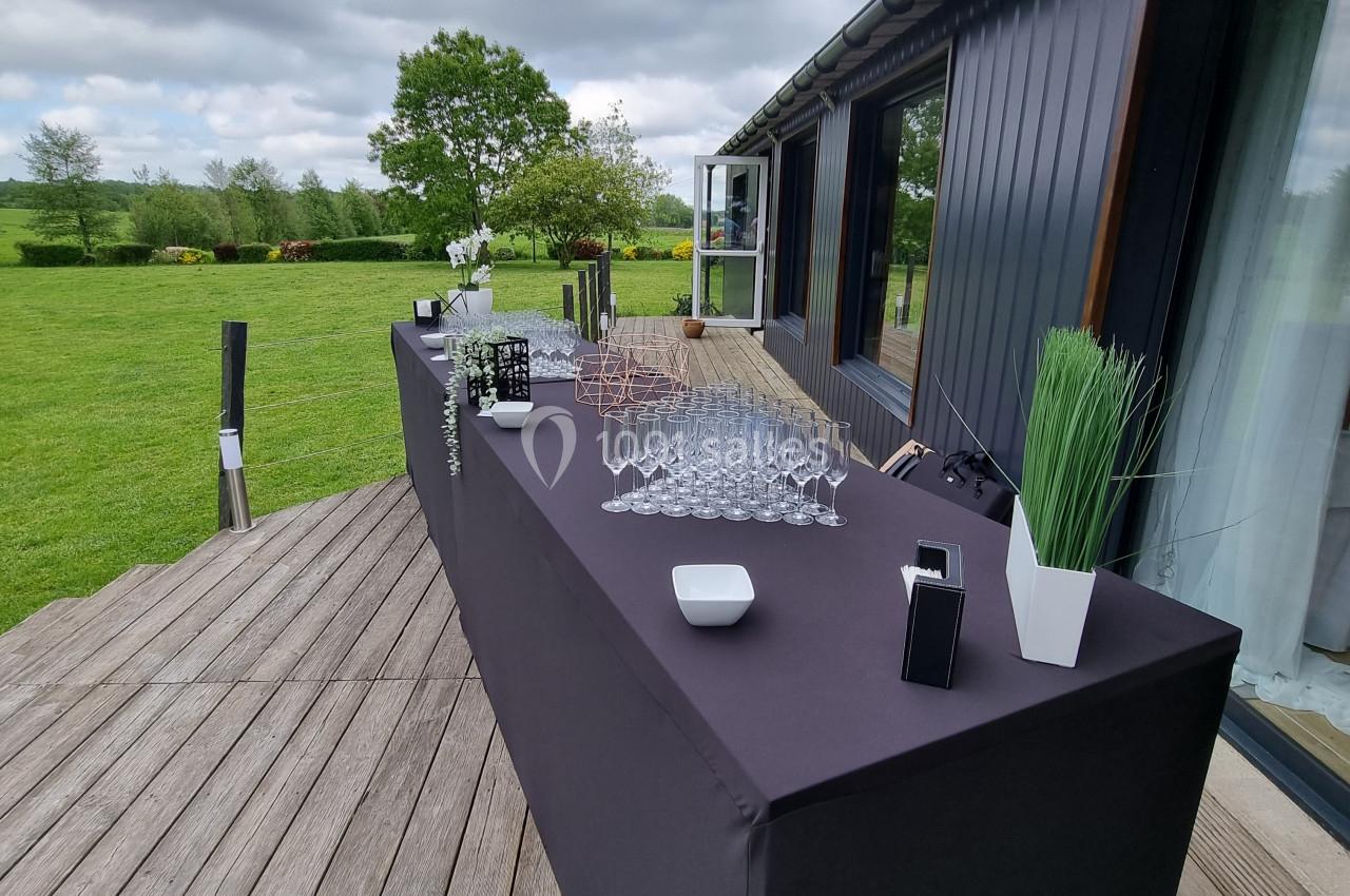 Table dressée en extérieur sur une terrasse en bois, avec verres, bouteilles et décorations, sous un ciel nuageux.