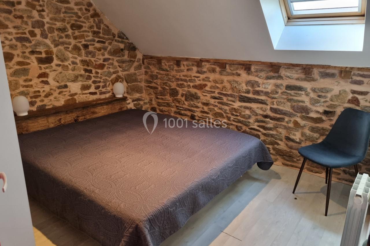 Chambre mansardée avec un lit double, mur en pierres apparentes, chaise noire et fenêtre de toit laissant entrer la lumière.