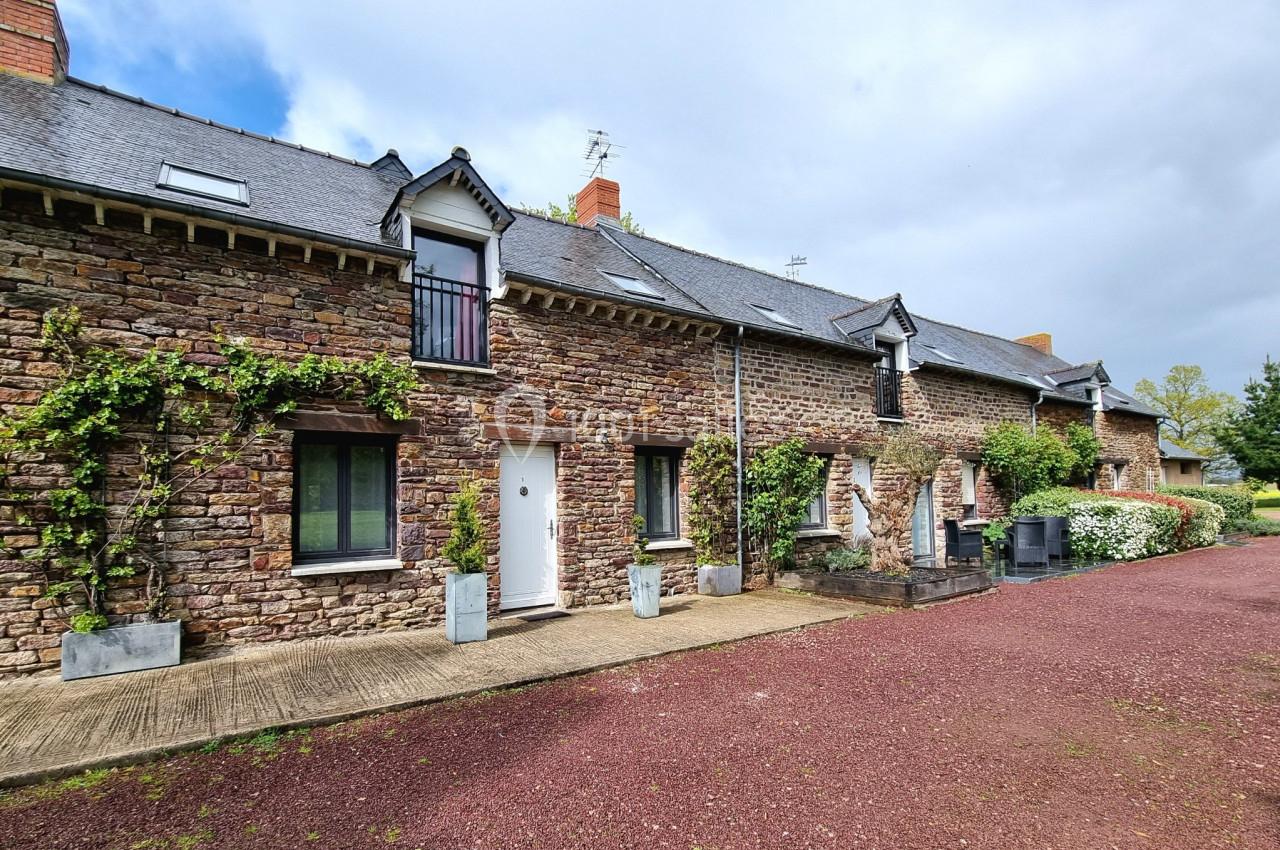 Façade en pierre d'une maison traditionnelle avec volets noirs, entourée de végétation et d'une allée en gravier rouge.