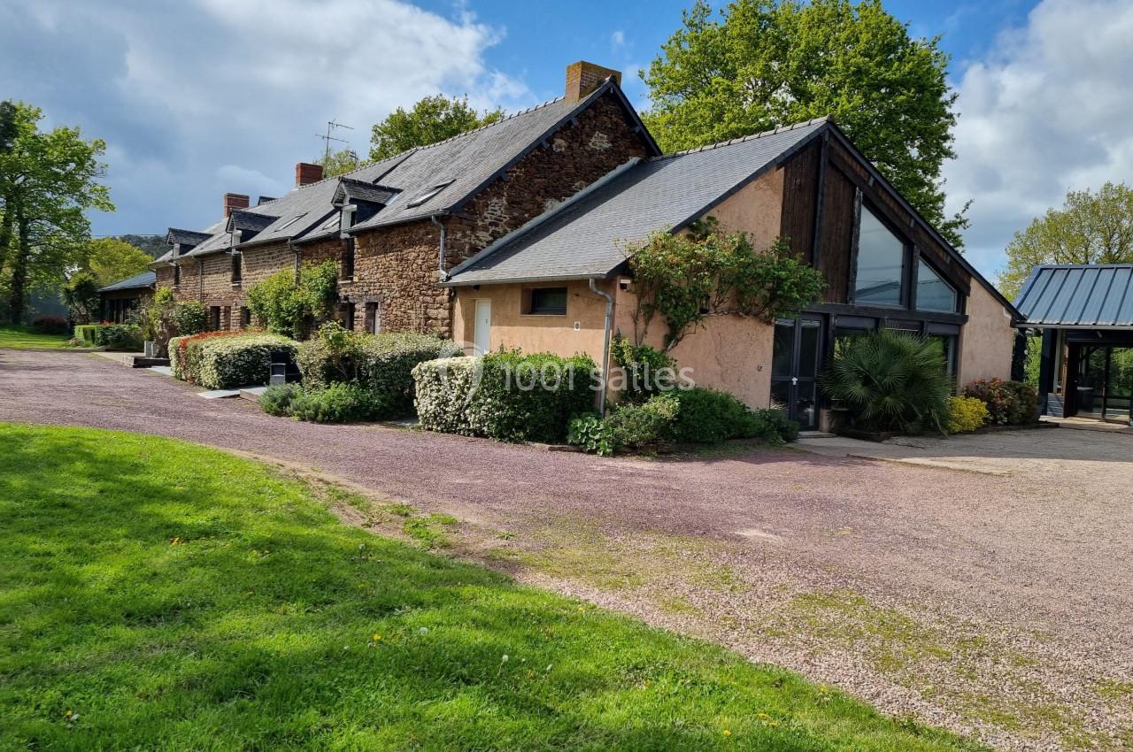 Vue d'un bâtiment en pierre et crépi avec toit en ardoise, entouré de végétation et d'une allée gravillonnée.