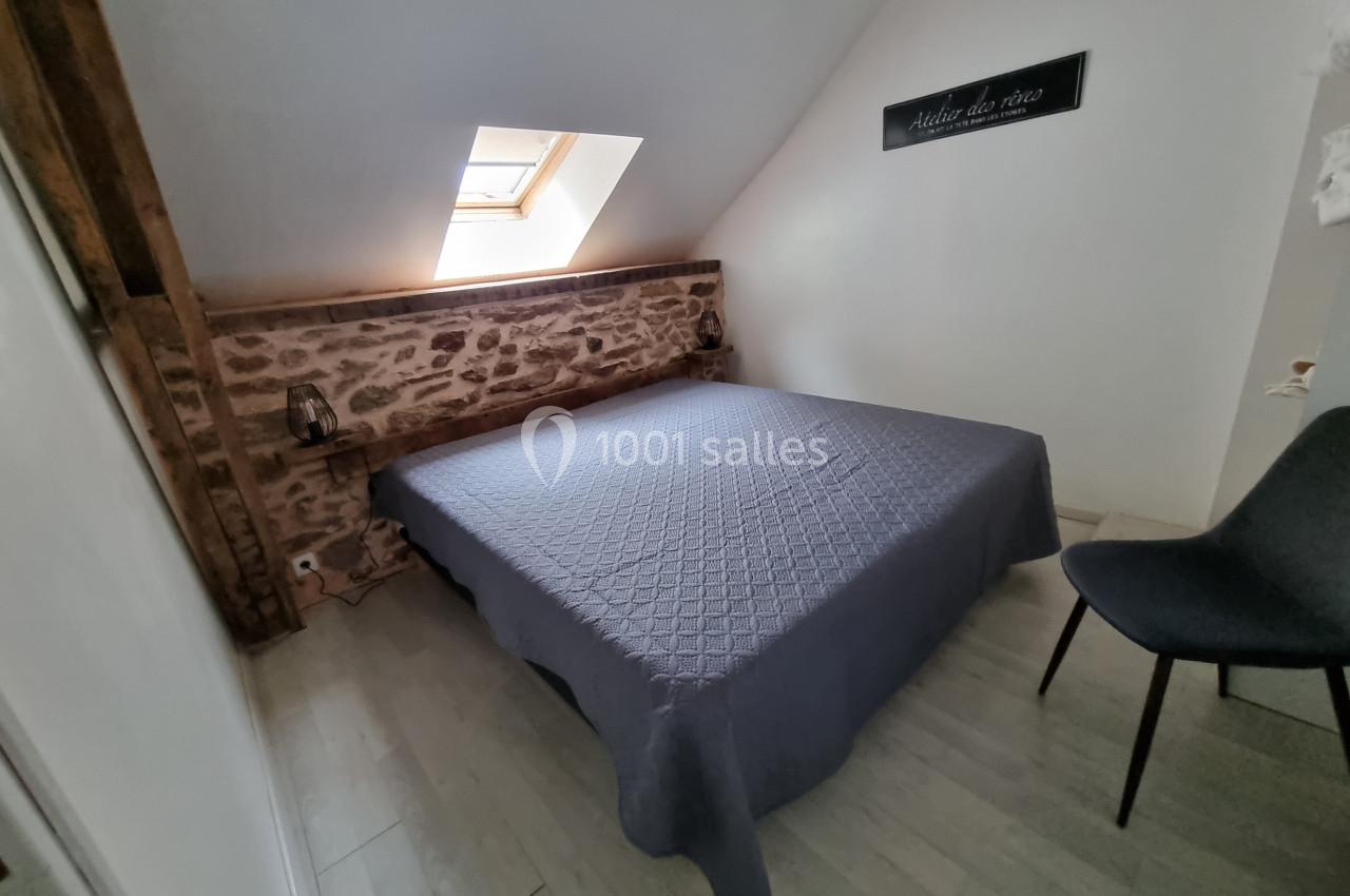 Chambre mansardée avec lit double, mur en pierre apparente, fenêtre de toit et chaise grise sur sol clair.