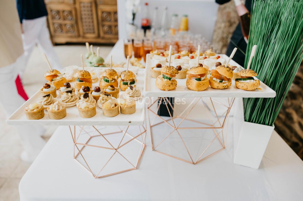 Plateaux de bouchées apéritives variées présentées sur des supports géométriques, avec des boissons en arrière-plan.