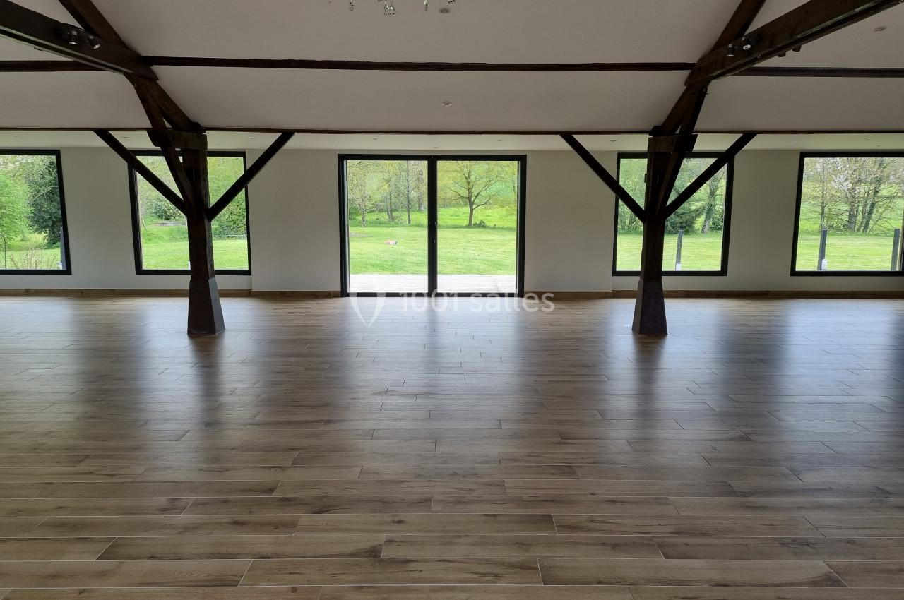 Salle vide avec parquet en bois, poutres apparentes et grandes baies vitrées donnant sur un espace vert.