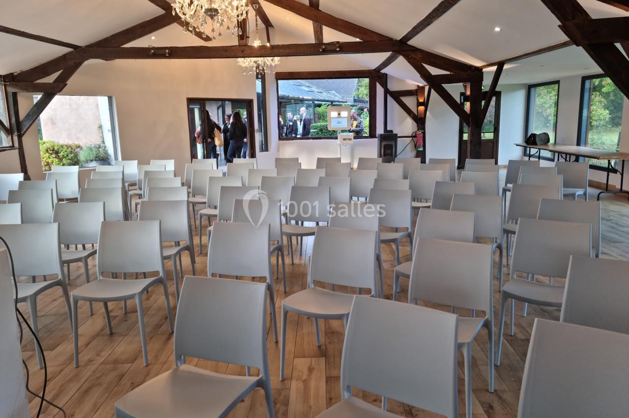 Salle lumineuse avec des rangées de chaises blanches alignées, poutres apparentes et lustre au plafond.