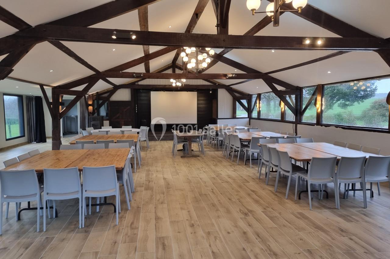 Salle spacieuse avec charpente en bois, tables et chaises disposées pour un événement, éclairage naturel et artificiel.