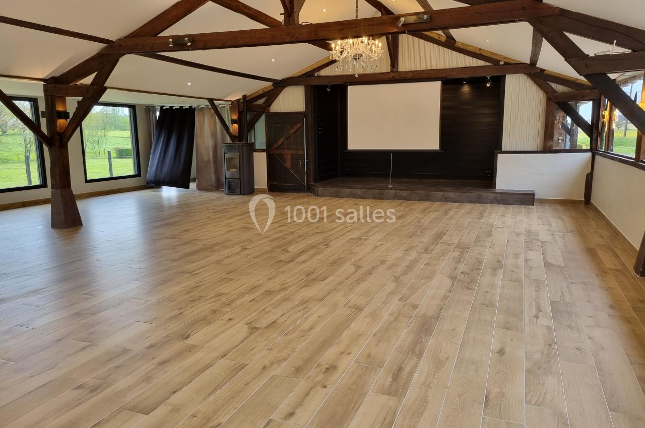 Salle spacieuse avec parquet en bois, poutres apparentes, scène surélevée et grandes fenêtres donnant sur un extérieur…
