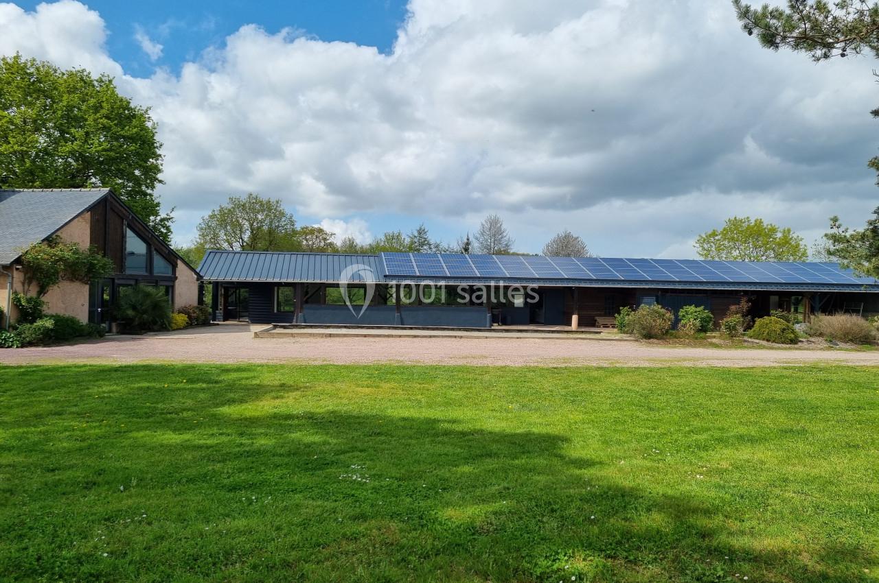 Bâtiment moderne avec toit équipé de panneaux solaires, entouré de verdure et sous un ciel partiellement nuageux.
