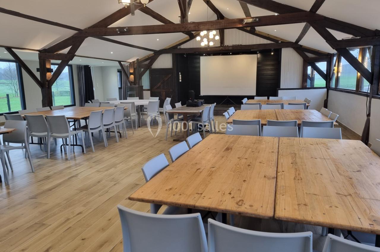 Salle lumineuse avec poutres apparentes, tables en bois, chaises modernes et écran de projection au fond.