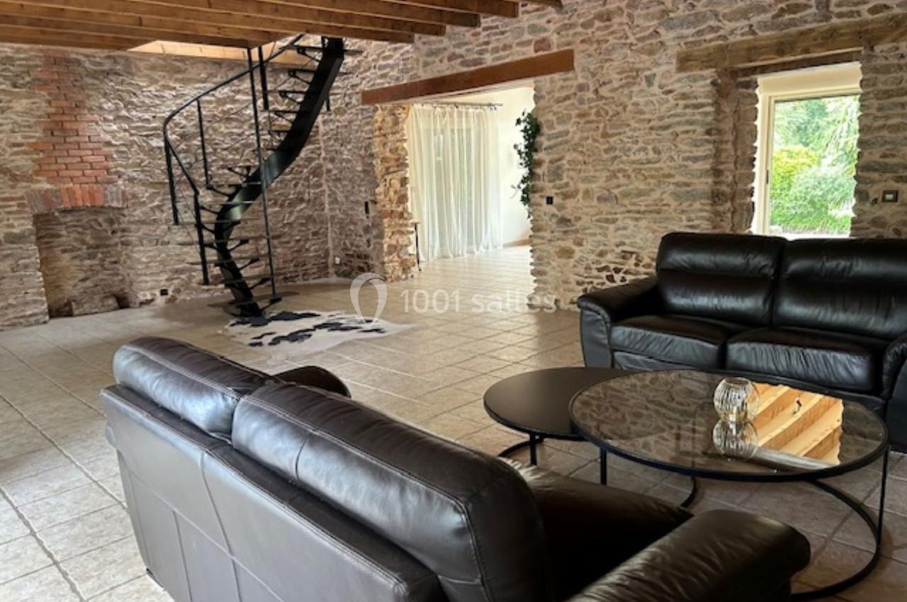 Salon spacieux avec murs en pierre apparente, canapé en cuir, table basse en verre et escalier en colimaçon.