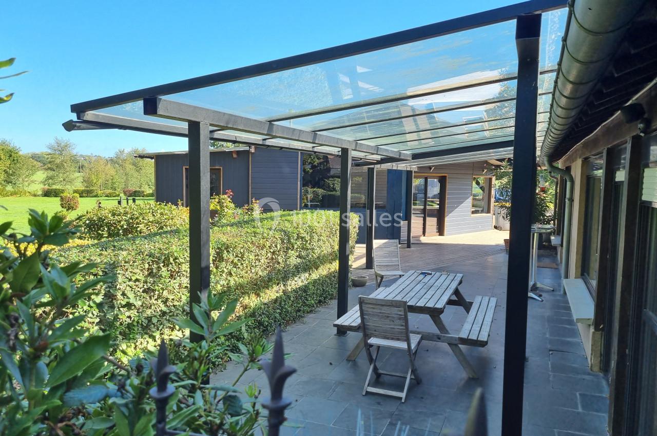 Terrasse extérieure avec toit en verre, mobilier en bois et vue sur un jardin verdoyant par une journée ensoleillée.