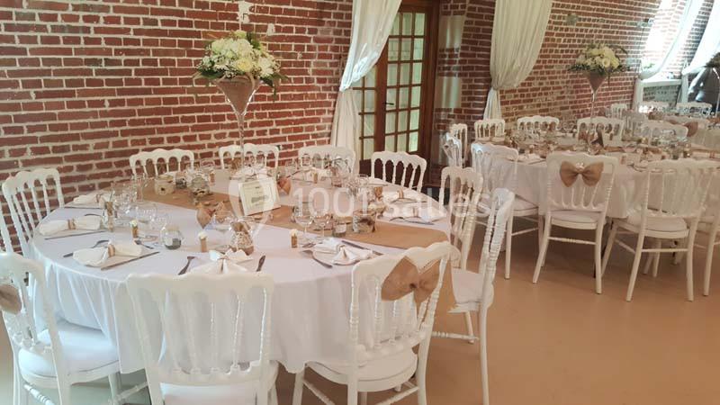 Salle décorée pour un événement avec tables rondes, nappes blanches, chaises assorties et centres de table fleuris.