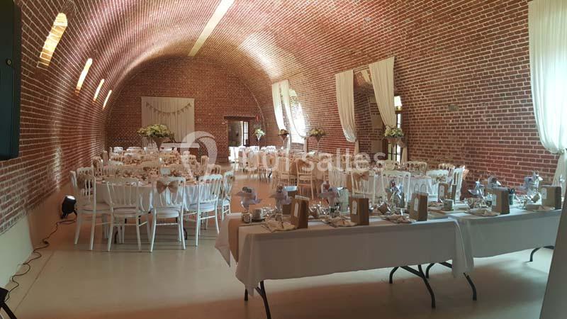 Salle voûtée en briques rouges aménagée pour un mariage, avec tables décorées et chaises blanches alignées.