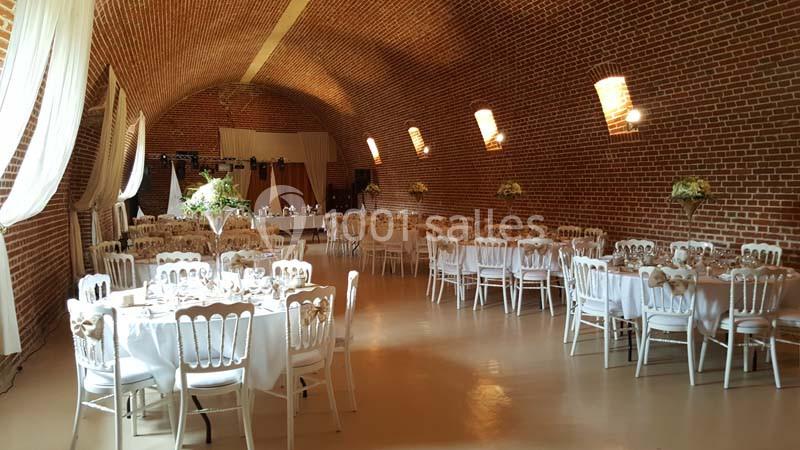 Salle de réception en briques rouges avec tables rondes dressées, chaises blanches et décorations florales.