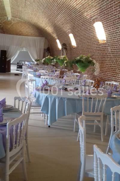 Salle voûtée en briques aménagée pour un événement, avec tables dressées, nappes bleues et chaises blanches.