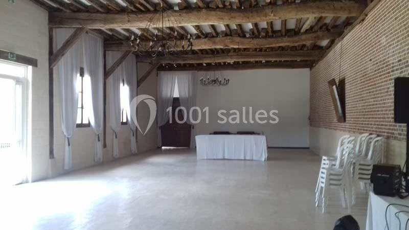 Salle vide avec murs en briques, poutres apparentes, rideaux blancs et une table recouverte d'une nappe blanche.