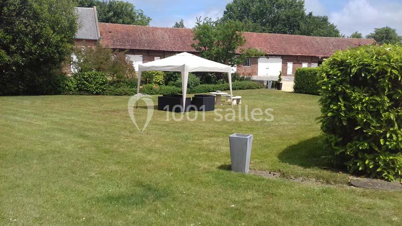 Tente blanche avec mobilier de jardin sur une pelouse, entourée de haies et d'un bâtiment en briques rouges.