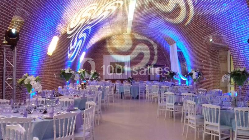 Salle voûtée en briques décorée pour un événement, avec tables dressées, nappes bleues et éclairage d'ambiance.