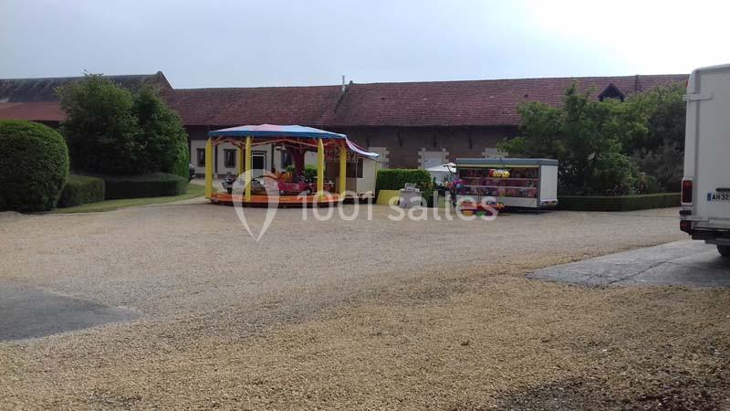 Un petit manège coloré et une remorque de stand sont installés sur une place en gravier devant des bâtiments ruraux.