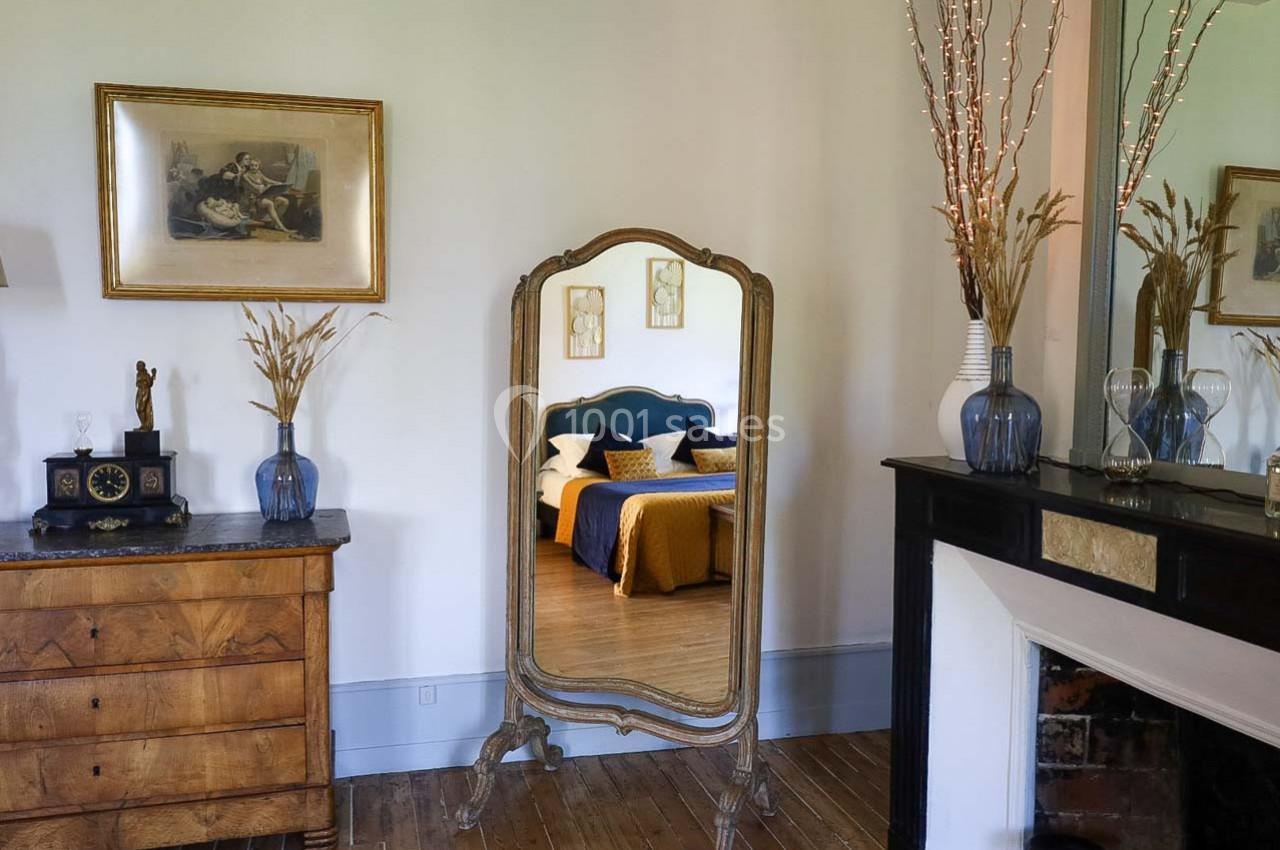 Miroir ancien sur pied reflétant un lit avec couvre-lit jaune, entouré de meubles et décorations classiques.