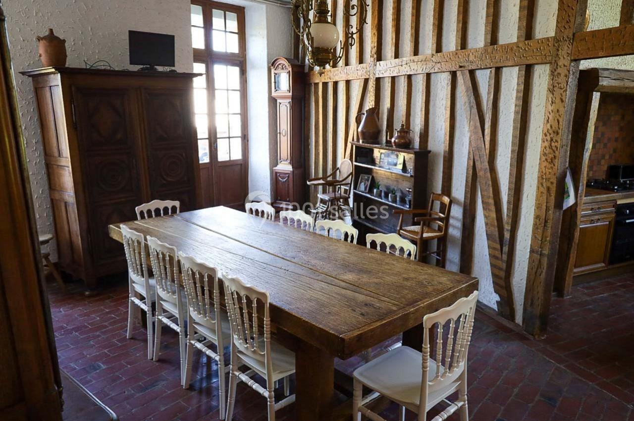 Salle à manger rustique avec une grande table en bois, des chaises blanches, des poutres apparentes et une cuisine en…