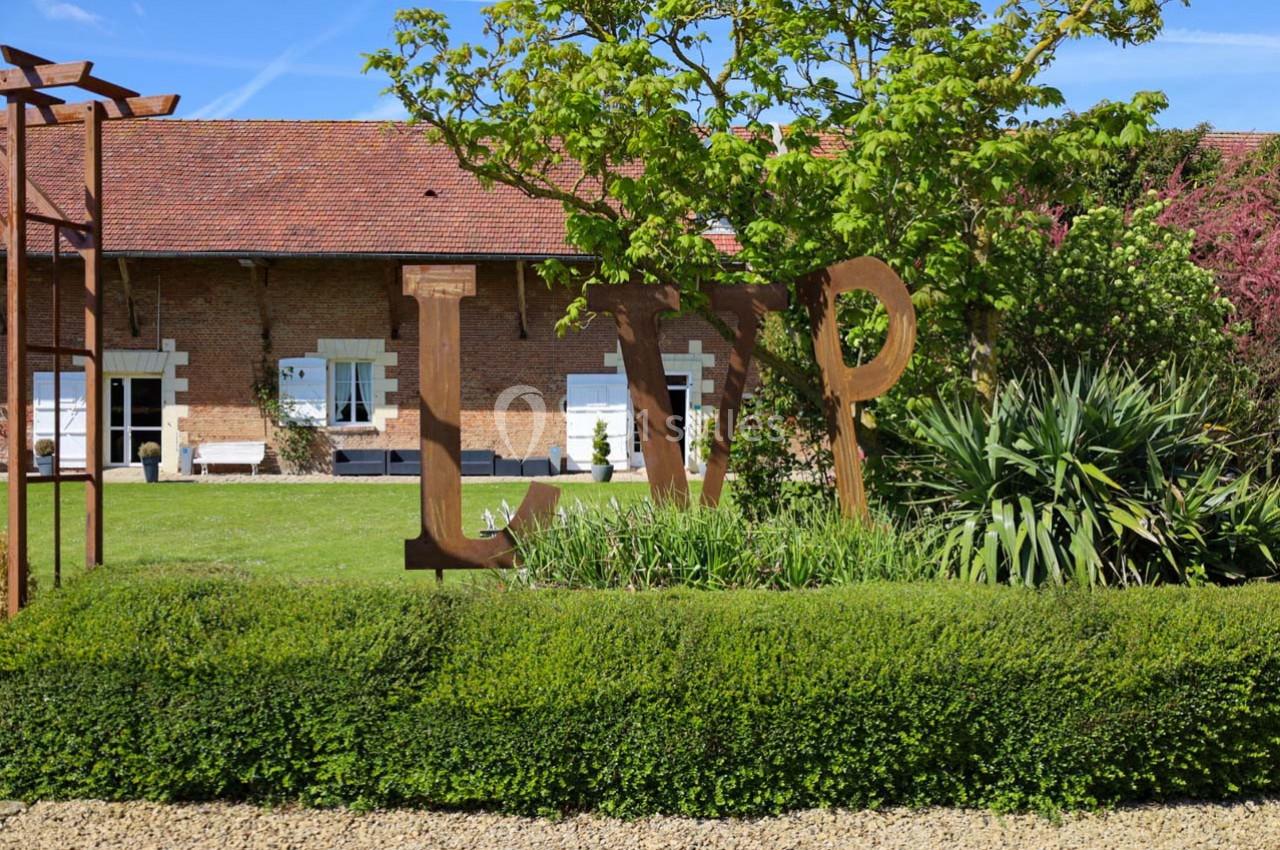 Jardin paysager avec haies taillées, sculptures métalliques et bâtiment en briques avec volets blancs en arrière-plan.