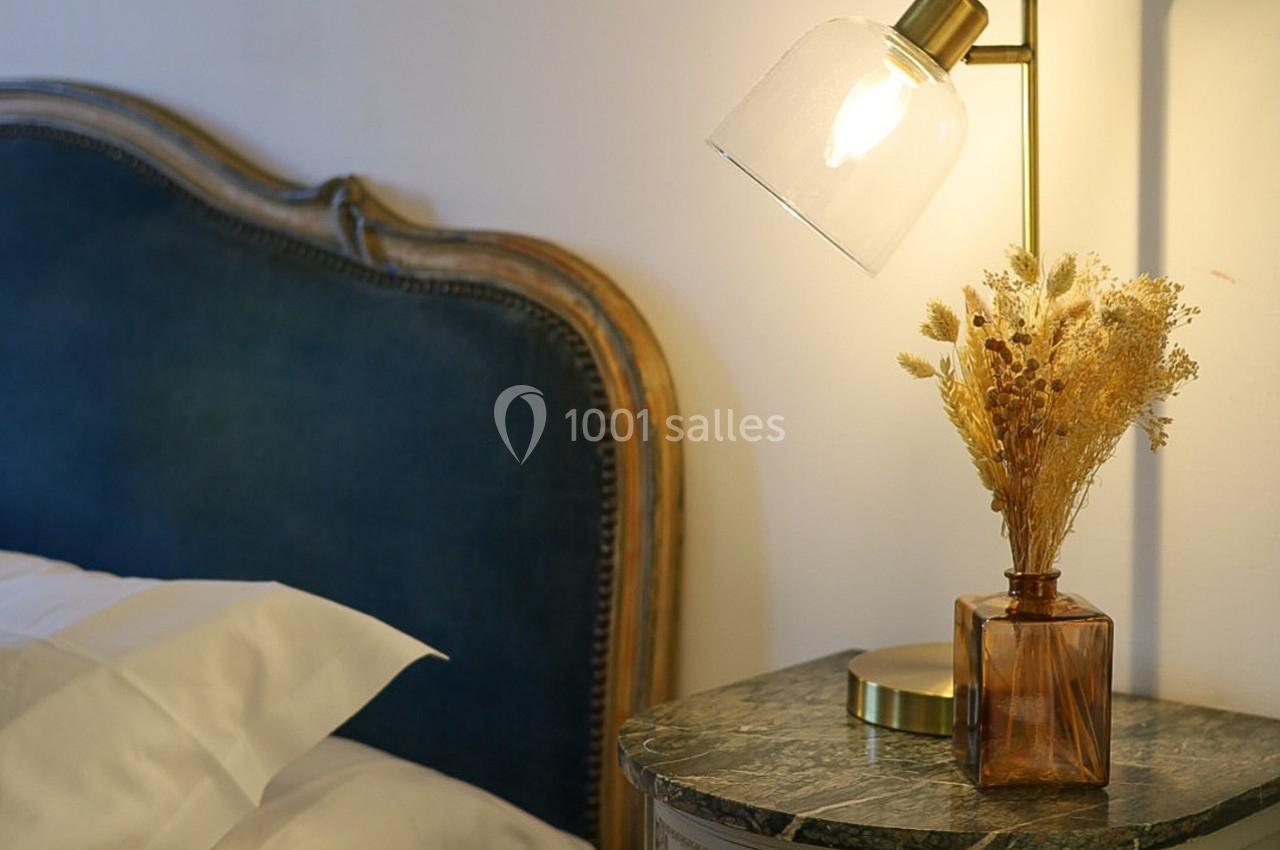 Lampe allumée sur une table de chevet en marbre avec un vase de fleurs séchées, près d'un lit avec tête de lit bleue.