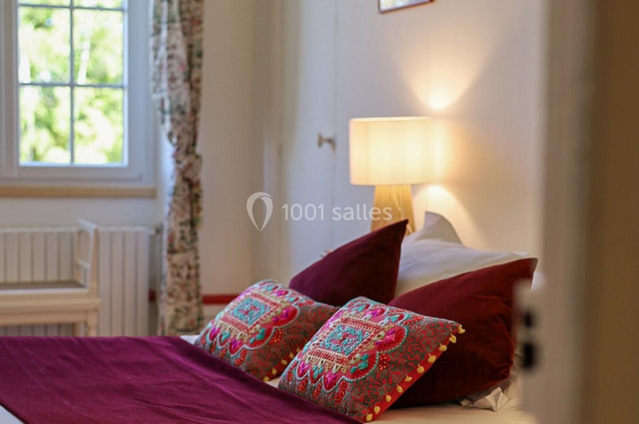 Lit avec coussins colorés et couverture bordeaux dans une chambre lumineuse avec lampe allumée et fenêtre donnant sur l…