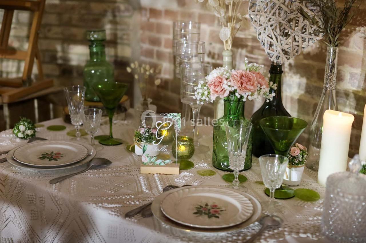 Table décorée avec vaisselle florale, verres en cristal, bougies et vases verts contenant des fleurs roses et blanches.