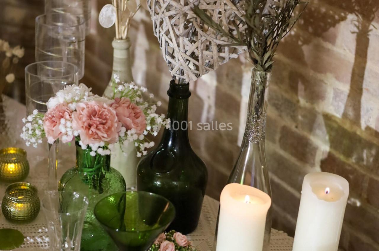 Décoration de table avec bougies allumées, fleurs roses, vases en verre vert et un cœur en osier devant un mur en briques.