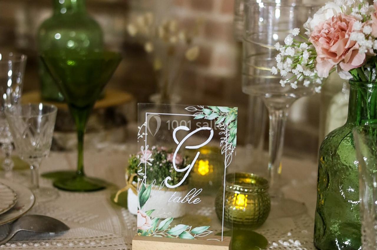 Carte de table décorative avec lettre ’G’, entourée de fleurs et verres sur une table élégamment dressée.