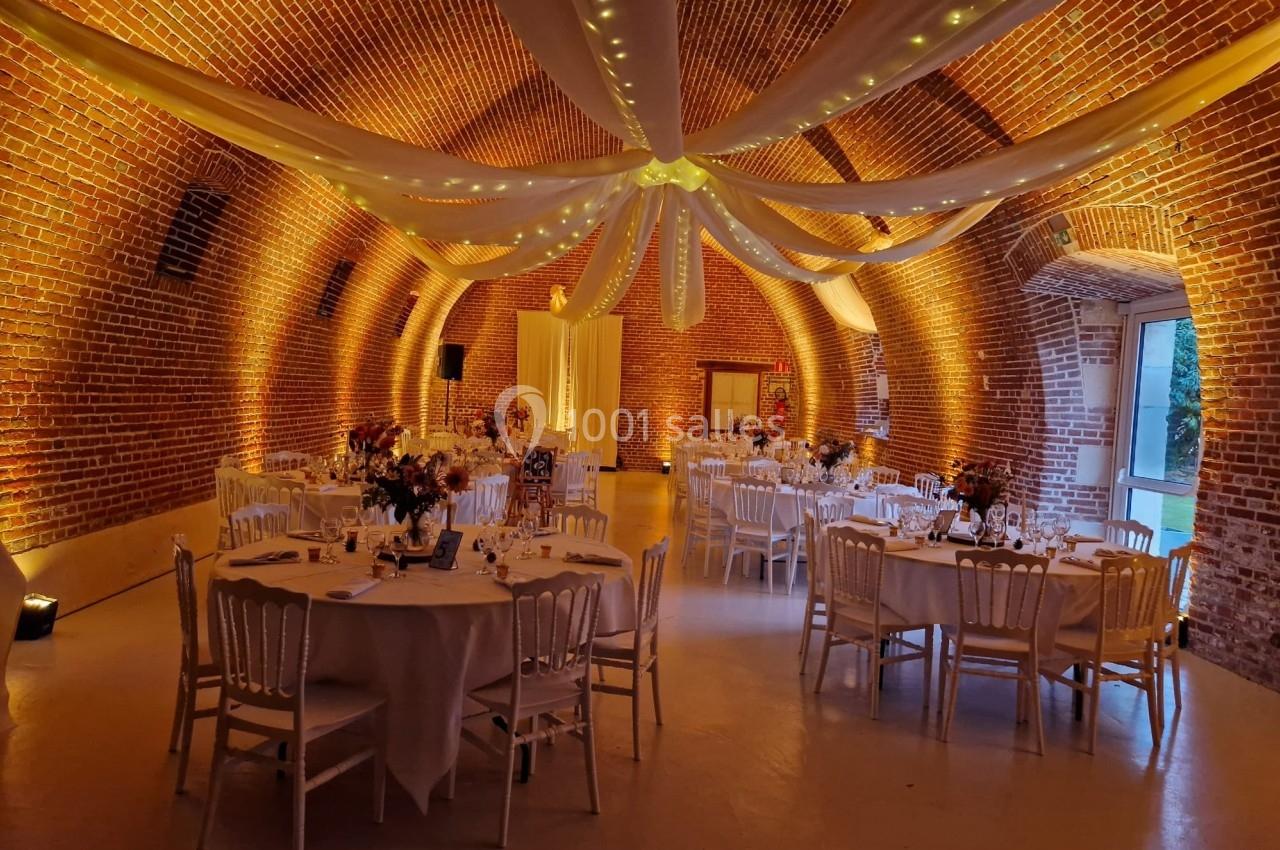 Salle voûtée en briques rouges décorée de guirlandes lumineuses et de tables rondes dressées pour un événement.