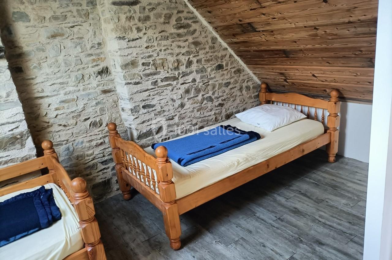 Deux lits simples en bois avec draps et couvertures bleues, dans une chambre mansardée aux murs en pierre et plafond en bois.