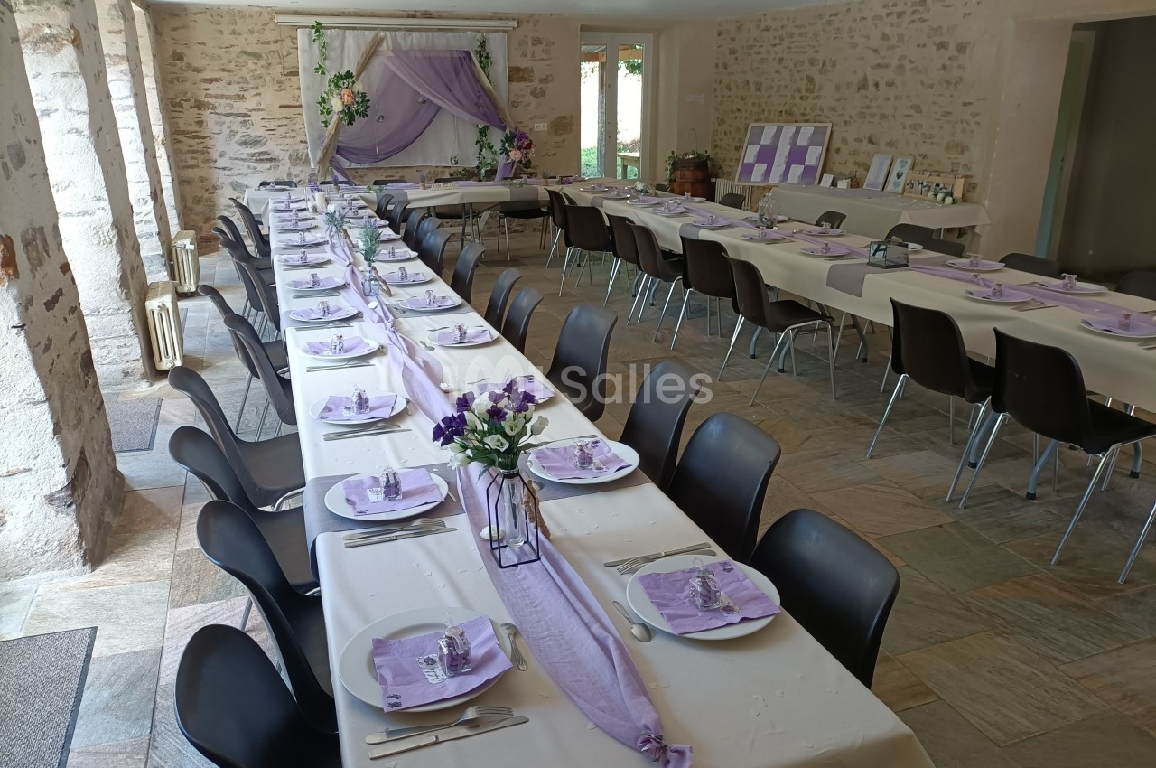 Salle décorée pour un événement, avec de longues tables dressées, nappes blanches et accents violets.
