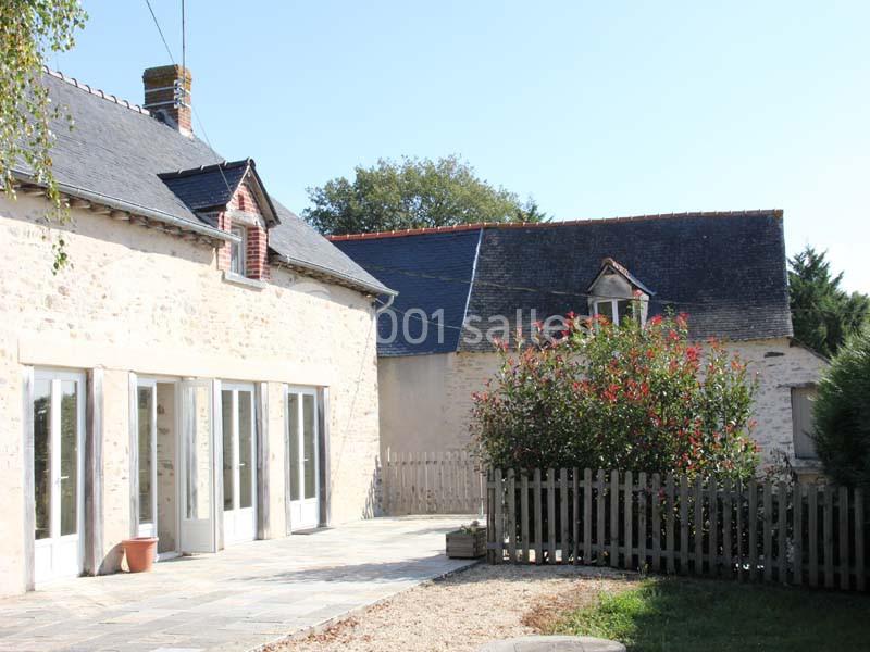 Façade en pierre d'une maison avec terrasse pavée, clôture en bois et arbuste fleuri sous un ciel dégagé.