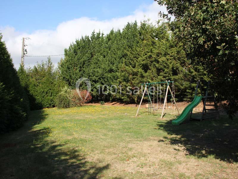 Jardin avec pelouse, balançoire en bois et toboggan vert, entouré d'arbres et arbustes sous un ciel partiellement nuageux.
