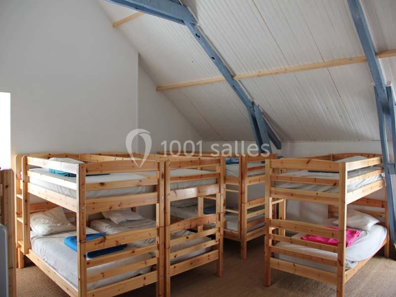 Dortoir avec plusieurs lits superposés en bois sous un plafond mansardé, éclairé par une lumière naturelle.