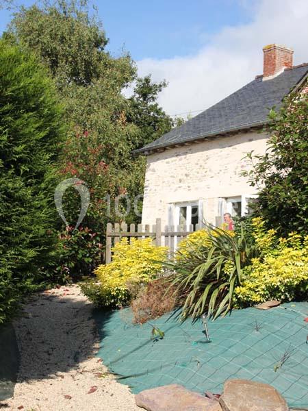 Maison en pierre avec toit en ardoise, entourée de végétation, arbustes et jardin aménagé avec graviers et plantes.